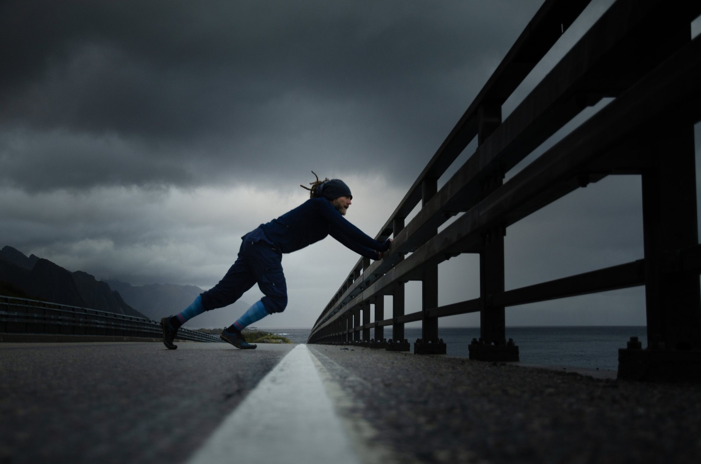 Un coureur en chaussettes bleues pousse de tout son corps contre la rambarde d’un pont, sous un ciel d’orage, face aux montagnes.